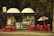 A small kiosk surrounded by trees featuring a variety of signs and advertisements is depicted. Bright red tables and chairs are positioned outside, some beneath a Coca Cola-branded umbrella. There are colorful posters and products displayed inside the kiosk.