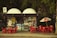 A small kiosk surrounded by trees featuring a variety of signs and advertisements is depicted. Bright red tables and chairs are positioned outside, some beneath a Coca Cola-branded umbrella. There are colorful posters and products displayed inside the kiosk.