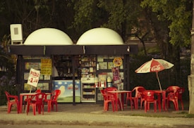 A small kiosk surrounded by trees featuring a variety of signs and advertisements is depicted. Bright red tables and chairs are positioned outside, some beneath a Coca Cola-branded umbrella. There are colorful posters and products displayed inside the kiosk.