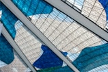 Intricate stained glass panels with blue, white, and green hues form an artistic pattern. The panels are intersected by white beams, lending a geometric framework to the fluid, colorful design. The glass features a mesh of thin black lines, creating an abstract and modern aesthetic.