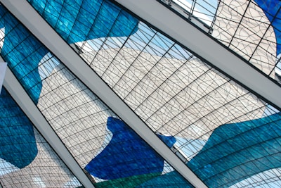 Intricate stained glass panels with blue, white, and green hues form an artistic pattern. The panels are intersected by white beams, lending a geometric framework to the fluid, colorful design. The glass features a mesh of thin black lines, creating an abstract and modern aesthetic.