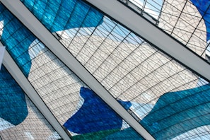 Intricate stained glass panels with blue, white, and green hues form an artistic pattern. The panels are intersected by white beams, lending a geometric framework to the fluid, colorful design. The glass features a mesh of thin black lines, creating an abstract and modern aesthetic.