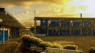A school building with blue accents is seen through a window with raindrops. The sky is partly cloudy with a golden hue from the setting or rising sun. The foreground includes a green lawn and a small fence, with rain adding a sense of calmness.
