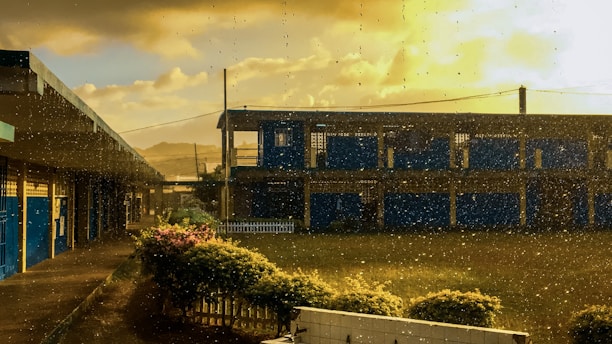 A school building with blue accents is seen through a window with raindrops. The sky is partly cloudy with a golden hue from the setting or rising sun. The foreground includes a green lawn and a small fence, with rain adding a sense of calmness.