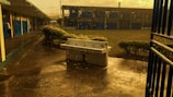 The image depicts an empty school courtyard during rainfall. There is a white tiled sink without water taps in the middle of the paved ground. Water droplets can be seen falling and splashing on the ground. To the left, there are blue doors and a covered walkway. In the background, there is a two-story building with numerous blue shutters and small bushes around.
