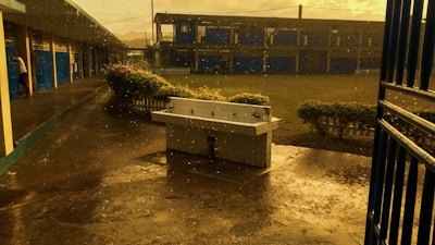 The image depicts an empty school courtyard during rainfall. There is a white tiled sink without water taps in the middle of the paved ground. Water droplets can be seen falling and splashing on the ground. To the left, there are blue doors and a covered walkway. In the background, there is a two-story building with numerous blue shutters and small bushes around.