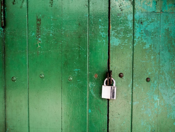 A weathered green wooden door with vertical panels, featuring a large silver padlock securing it. The door shows signs of age with peeling paint and visible wear. The padlock is attached to a metal latch.