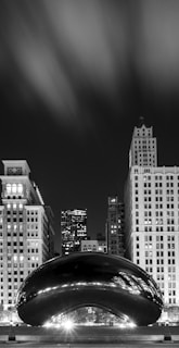 A black and white cityscape featuring a modern, reflective sculpture set against tall, illuminated buildings. The sky appears cloudy, enhancing the dramatic contrast in the scene.