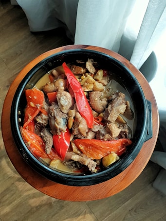 A black pot is filled with a hearty stew featuring chunks of meat, sliced red bell peppers, and what appear to be pieces of eggplant. The pot rests on a wooden surface with a warm, sunlit atmosphere.