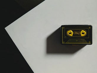 A vintage cassette tape with a black and yellow design sits on a flat surface casting a shadow. The tape is decorated with illustrations of two reels labeled with yellow text. It's positioned on a geometric, possibly triangular, section with contrasting shadows.