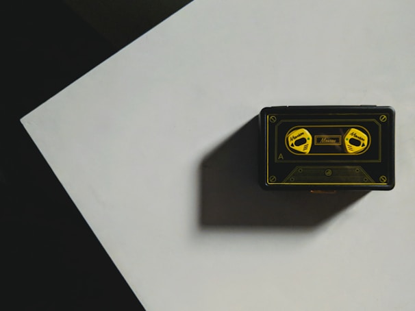A vintage cassette tape with a black and yellow design sits on a flat surface casting a shadow. The tape is decorated with illustrations of two reels labeled with yellow text. It's positioned on a geometric, possibly triangular, section with contrasting shadows.