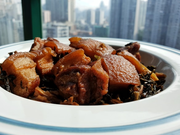 A plate of cooked pork with a rich glaze, mixed with vegetables, is placed on a white dish with a blue rim. The meal is in focus with a blurred urban skyline in the background, suggesting a city setting.