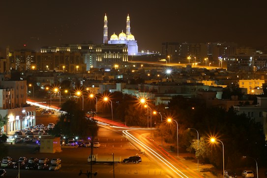 The image captures a night scene of an illuminated mosque with multiple domes and minarets standing prominently in the background. The cityscape around the mosque is bustling with street lights and light trails from moving vehicles on the roads, indicating active traffic. Buildings of various sizes and structures fill the midground and foreground, with some showing dimly lit interiors. Trees and parked vehicles are also visible, adding to the nighttime urban ambiance.