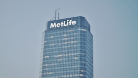 A tall, modern skyscraper with a glass facade and the word 'MetLife' displayed prominently near the top. The building has multiple floors with lights visible in some windows. The sky is clear and gives a serene backdrop to the structure.