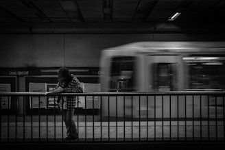 A black and white image showcasing a person standing at a subway station platform, leaning slightly over a railing while reading a piece of paper. In the background, a train is moving past at high speed, appearing blurred due to motion. The setting indicates an urban transit environment, with 'Civic Center' signage visible on the wall.