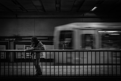 A black and white image showcasing a person standing at a subway station platform, leaning slightly over a railing while reading a piece of paper. In the background, a train is moving past at high speed, appearing blurred due to motion. The setting indicates an urban transit environment, with 'Civic Center' signage visible on the wall.