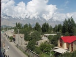 A residential area surrounded by trees with mountains in the background. The street is lined with small buildings and a few pedestrians are walking along the road. A prominent house with a red roof stands out on the right side.
