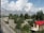 A residential area surrounded by trees with mountains in the background. The street is lined with small buildings and a few pedestrians are walking along the road. A prominent house with a red roof stands out on the right side.