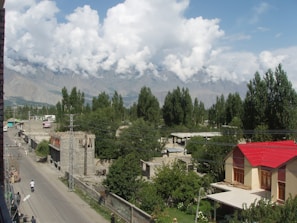 A residential area surrounded by trees with mountains in the background. The street is lined with small buildings and a few pedestrians are walking along the road. A prominent house with a red roof stands out on the right side.