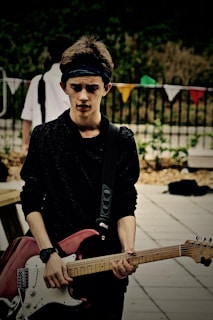 A young person is holding an electric guitar outdoors with a serious expression. The background includes blurred greenery and colorful triangular flags. The individual is wearing a dark sweater and a headband.