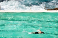 A swimmer in motion in a turquoise swimming pool with ocean waves crashing in the background. A white concrete barrier separates the pool from the sea, with safety chains attached to poles along the edge. The swimmer wears black shorts and the scene captures a blend of artificial and natural aquatic environments.