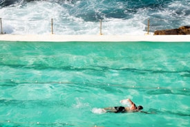 A swimmer in motion in a turquoise swimming pool with ocean waves crashing in the background. A white concrete barrier separates the pool from the sea, with safety chains attached to poles along the edge. The swimmer wears black shorts and the scene captures a blend of artificial and natural aquatic environments.