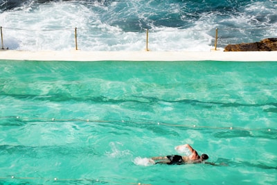 A swimmer in motion in a turquoise swimming pool with ocean waves crashing in the background. A white concrete barrier separates the pool from the sea, with safety chains attached to poles along the edge. The swimmer wears black shorts and the scene captures a blend of artificial and natural aquatic environments.