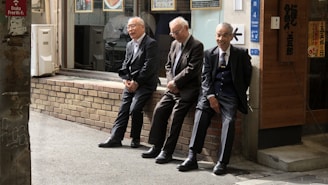 Three elderly men are dressed in suits, sitting on a low brick wall in an urban setting. They seem to be engaged in a casual and light-hearted conversation, with one of them laughing. The location appears to be near a traditional shop with Japanese signage, and the overall atmosphere is relaxed.