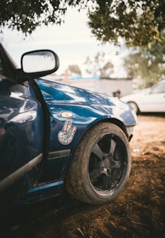 A close-up of the front side of a blue car with a decorative sticker on the fender. The vehicle has a large black wheel and is parked on a dirt surface, partially shaded by tree branches above. Another vehicle is visible in the background, slightly out of focus.