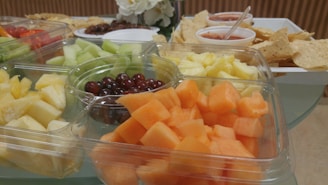A selection of fresh-cut fruits including cantaloupe, honeydew, pineapple, and grapes is arranged in a clear plastic tray. In the background, tortilla chips with salsa and what appear to be cherry tomatoes on another tray can be seen. A white flower arrangement is also partially visible.