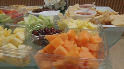 A selection of fresh-cut fruits including cantaloupe, honeydew, pineapple, and grapes is arranged in a clear plastic tray. In the background, tortilla chips with salsa and what appear to be cherry tomatoes on another tray can be seen. A white flower arrangement is also partially visible.
