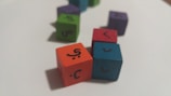 Colored wooden cubes with Arabic letters on them are scattered on a white surface. The cubes are in different colors such as orange, blue, red, green, and purple, arranged at different angles.