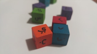 Colored wooden cubes with Arabic letters on them are scattered on a white surface. The cubes are in different colors such as orange, blue, red, green, and purple, arranged at different angles.