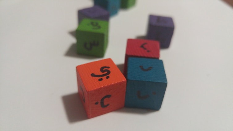 Colored wooden cubes with Arabic letters on them are scattered on a white surface. The cubes are in different colors such as orange, blue, red, green, and purple, arranged at different angles.