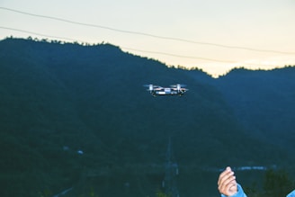 A small drone is flying in front of a mountainous landscape, captured during early evening. In the foreground, a person's hand is visible, possibly operating the drone. The background consists of a vast expanse of hills or mountains, with a muted sky due to the setting sun.