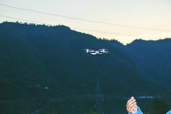 A small drone is flying in front of a mountainous landscape, captured during early evening. In the foreground, a person's hand is visible, possibly operating the drone. The background consists of a vast expanse of hills or mountains, with a muted sky due to the setting sun.