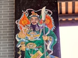 An artistic depiction of a warrior figure, adorned in ornate armor and vibrant robes. The figure is holding a large polearm weapon, wearing an elaborate headdress, and has facial hair. The background consists of architectural elements, possibly part of a traditional structure.