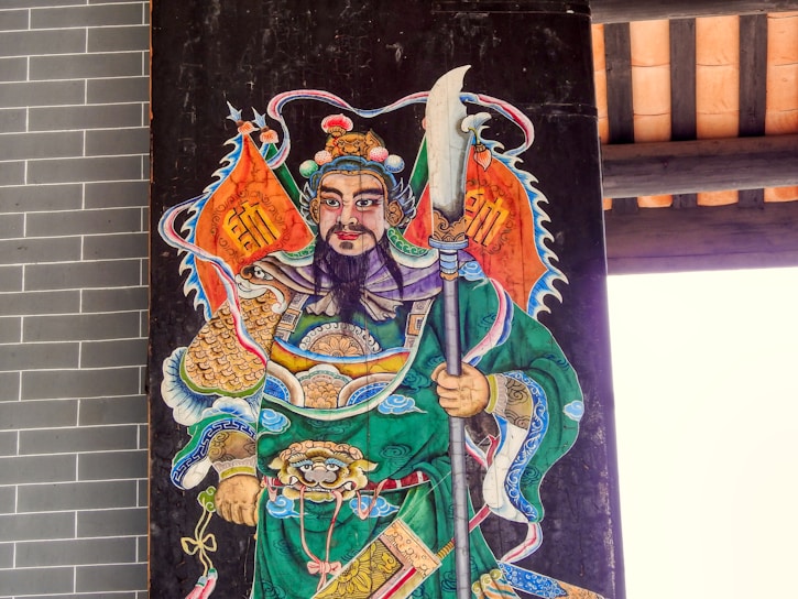 An artistic depiction of a warrior figure, adorned in ornate armor and vibrant robes. The figure is holding a large polearm weapon, wearing an elaborate headdress, and has facial hair. The background consists of architectural elements, possibly part of a traditional structure.