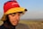 A person wearing a colorful hat with the word 'Colombia' on it stands outdoors in a natural setting. The hat is red, yellow, and blue, and the person has a beard. The background is a blurred landscape under a clear sky with soft lighting, suggesting either early morning or late afternoon.