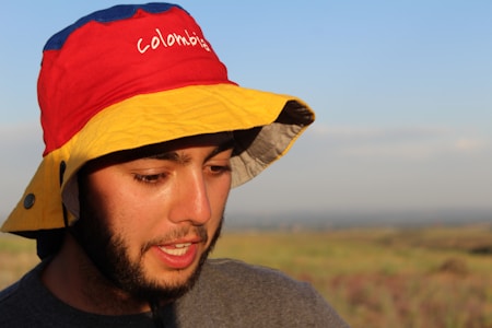 A person wearing a colorful hat with the word 'Colombia' on it stands outdoors in a natural setting. The hat is red, yellow, and blue, and the person has a beard. The background is a blurred landscape under a clear sky with soft lighting, suggesting either early morning or late afternoon.
