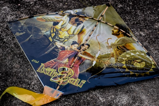 A kite featuring a dramatic scene with characters and weaponry, possibly from a historical or fantasy-themed movie. The kite showcases images of warriors with armor and weapons, including a man holding a bow and arrow. The background is a textured dark surface.