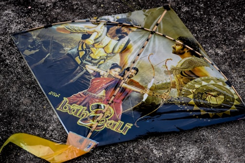 A kite featuring a dramatic scene with characters and weaponry, possibly from a historical or fantasy-themed movie. The kite showcases images of warriors with armor and weapons, including a man holding a bow and arrow. The background is a textured dark surface.