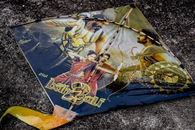 A kite featuring a dramatic scene with characters and weaponry, possibly from a historical or fantasy-themed movie. The kite showcases images of warriors with armor and weapons, including a man holding a bow and arrow. The background is a textured dark surface.