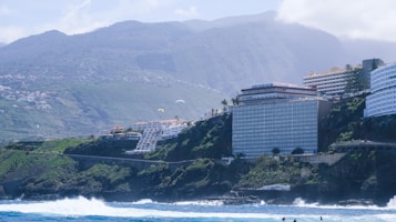 A coastal landscape featuring large buildings perched on cliffs overlooking the ocean. The backdrop consists of lush green hills and tall mountains partially covered by clouds. The sea is vibrant and blue, showing waves crashing against the rocky shoreline. Paragliders can be seen in the sky above the cliffs.