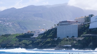 A coastal landscape featuring large buildings perched on cliffs overlooking the ocean. The backdrop consists of lush green hills and tall mountains partially covered by clouds. The sea is vibrant and blue, showing waves crashing against the rocky shoreline. Paragliders can be seen in the sky above the cliffs.