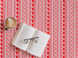 A patterned red and white tablecloth with a Nordic-inspired design featuring geometric shapes and reindeer motifs. Resting on the tablecloth are an open book with its pages slightly fanned, a pair of black-framed eyeglasses placed across the pages, and a cup of coffee with a latte art leaf pattern on its frothy surface.