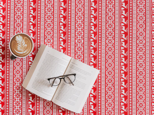 A patterned red and white tablecloth with a Nordic-inspired design featuring geometric shapes and reindeer motifs. Resting on the tablecloth are an open book with its pages slightly fanned, a pair of black-framed eyeglasses placed across the pages, and a cup of coffee with a latte art leaf pattern on its frothy surface.