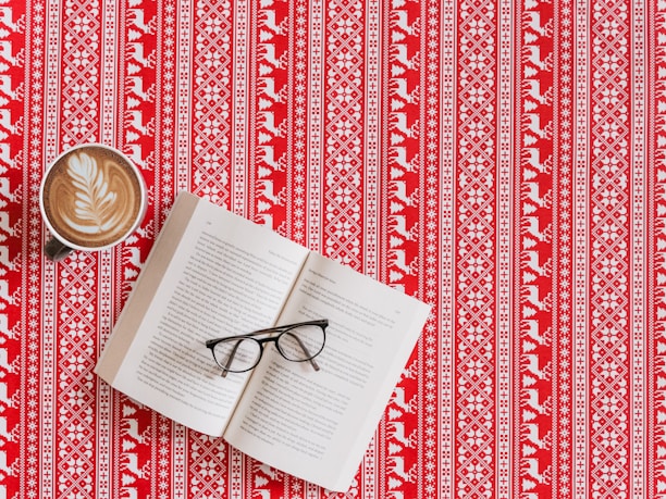 A patterned red and white tablecloth with a Nordic-inspired design featuring geometric shapes and reindeer motifs. Resting on the tablecloth are an open book with its pages slightly fanned, a pair of black-framed eyeglasses placed across the pages, and a cup of coffee with a latte art leaf pattern on its frothy surface.