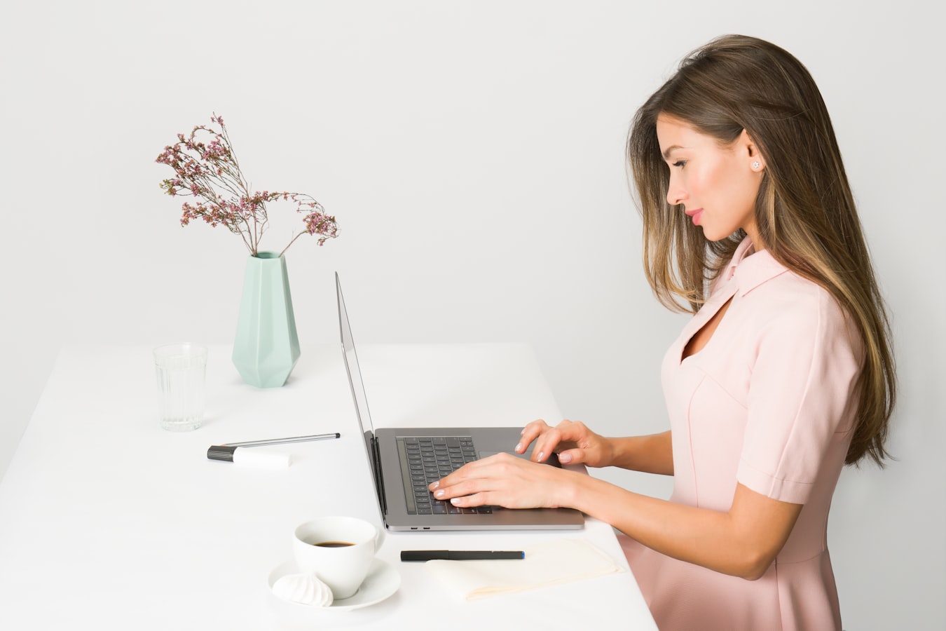 Woman with long brown hair wearing a pink dress typing on a laptop with cup of coffee and vase of flowers nearby