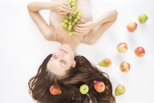 woman lying on white flooring holding grapes beside apple fruits